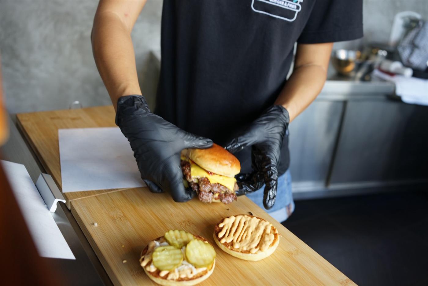Cook assembling a smashburger over sauced buns with pickles on a wooden board